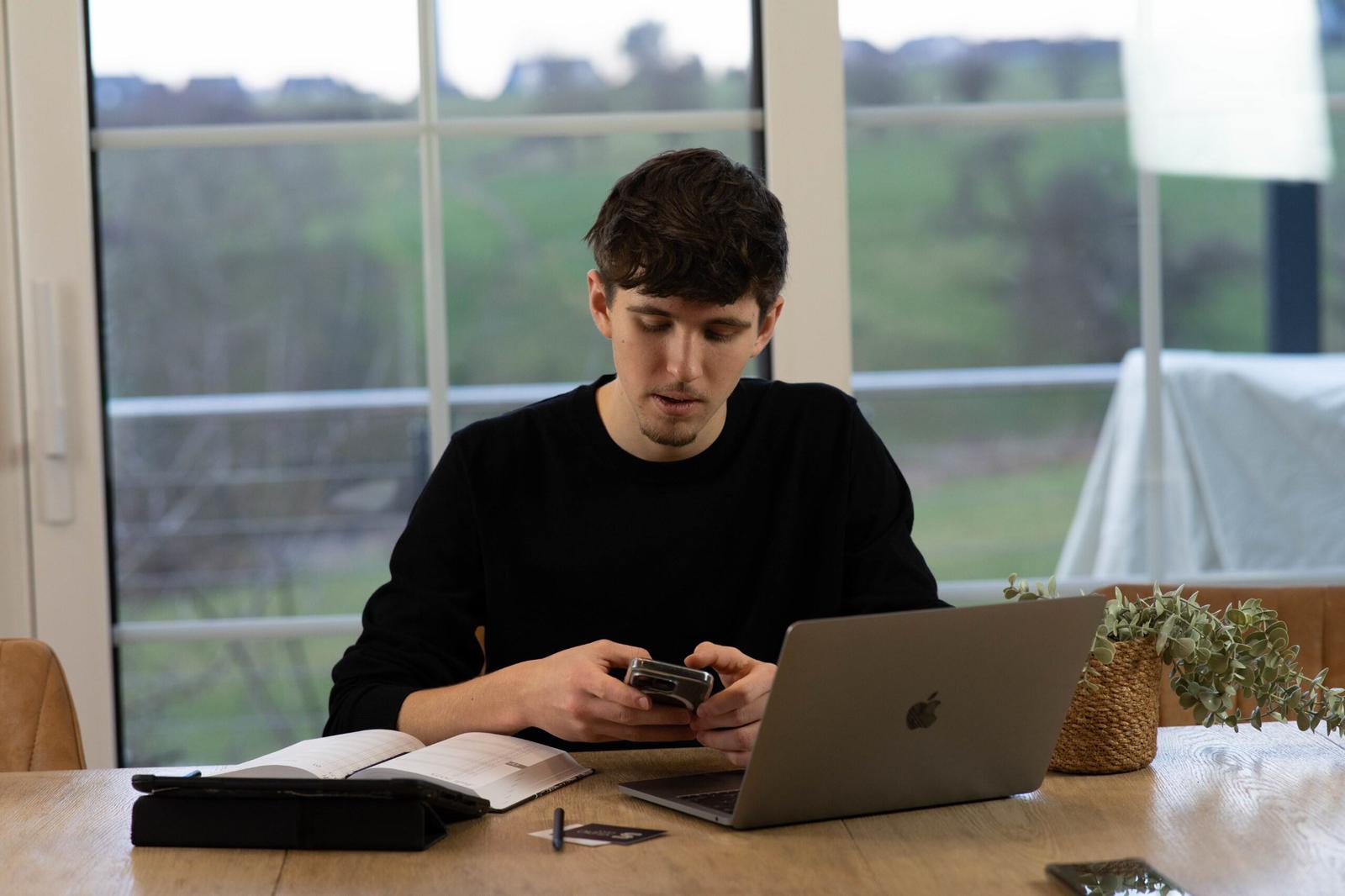 Person sitzt an einem Holztisch vor einem Laptop, hält ein Smartphone in den Händen und arbeitet zwischen Notizbuch, Karten und einer Pflanze; im Hintergrund große Fenster mit Blick ins Grüne.